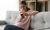 Mujer de jersey rosa en su sofá gris mirando su teléfono y sonriendo